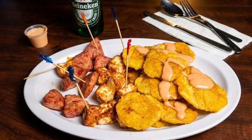 A plate of fried plantains with sauce, fried cheese, and sausage, with a Heineken bottle.