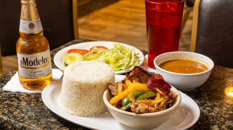 A Modelo beer, rice, meat with peppers, salad, soup, and a red drink on a table.