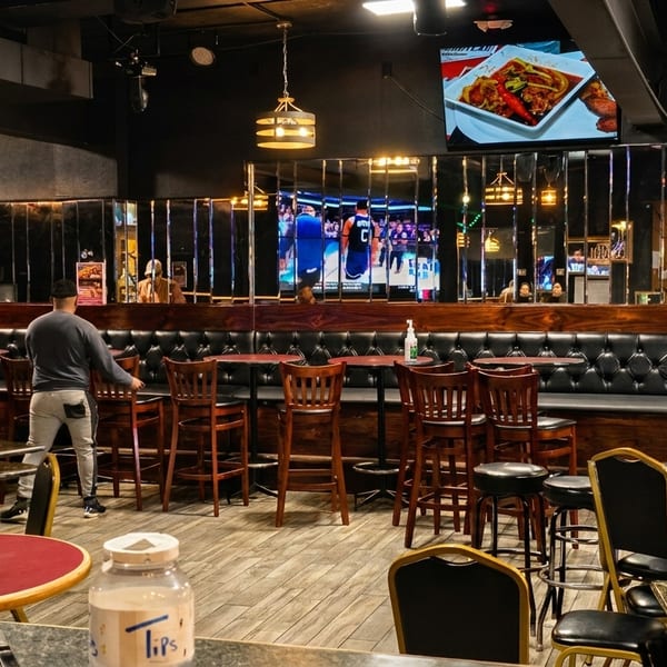 Two men arrange chairs and tables in a restaurant with dark decor, TVs, and a Corona Extra cutout.