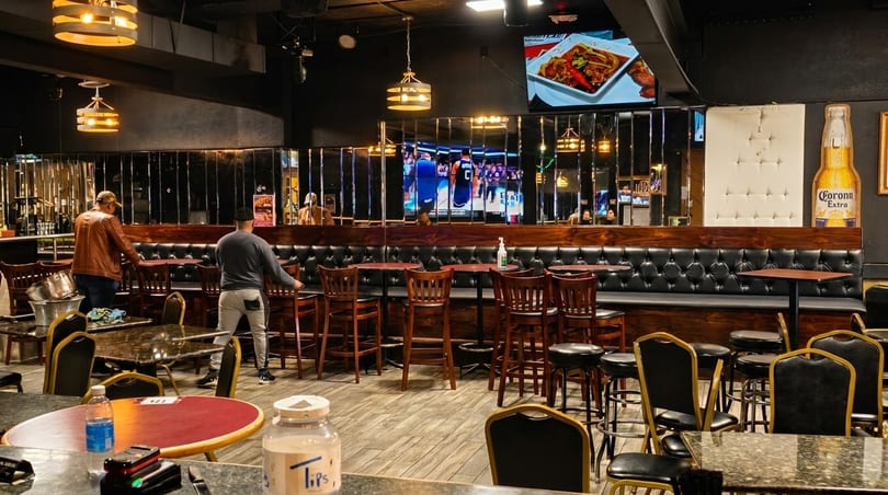 Two men arrange chairs and tables in a restaurant with dark decor, TVs, and a Corona Extra cutout.