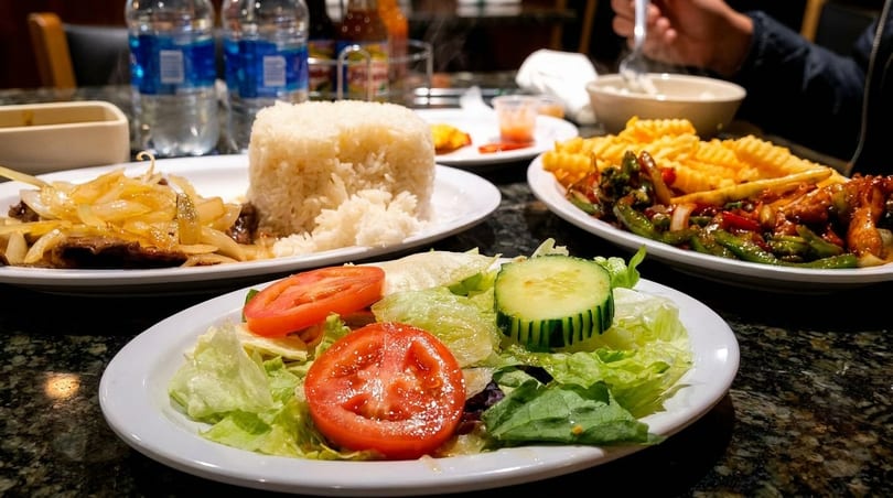 Plates of stir-fried beef and onions, rice, salad, and waffle fries on a dark table.
